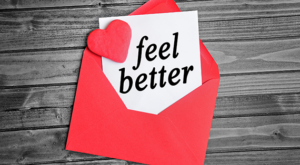 RED ENVELOPE WITH FEEL BETTER NOTE AND RED HEART ON A WOODEN PATTERN BACKGROUND
